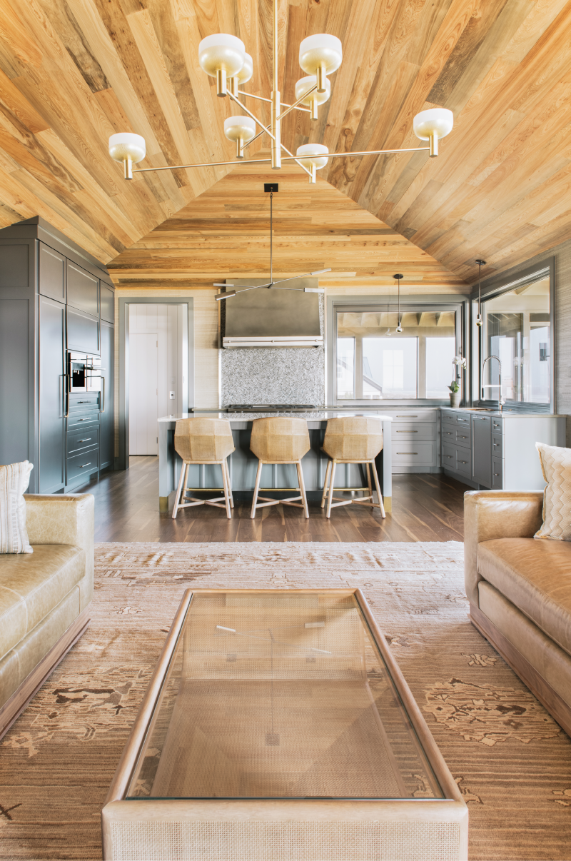Golden Hues: In the open-plan living/kitchen space, the ceiling of honey-colored, hand-placed sunken cypress planks contrasts beautifully with the custom cabinetry painted in “Down Pipe” by Farrow &amp; Ball. A vibrant tile backsplash, clad in “Hip Herringbone Detroit Blues” by Artistic Tile, sits under a custom exhaust hood made by Handcrafted Metal. Three McGuire rattan stools at the island offer occupants a view through the sliding window pass-through to the dining porch.