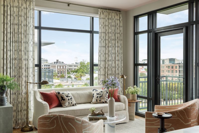 An Inviting Aerie: The condo’s open-plan living space encompasses a sitting room, breakfast nook, and kitchen. Low-slung furniture allows for unimpeded views, and the simple barrel shape of two Baker Furniture chairs covered in a Kelly Wearstler for Lee Jofa fabric in the sitting room adds warmth and style. Their sleek bronze legs and a vintage 1970s coffee table contrast with the simple white sofa and the pretty patterned drapes from Quadrille.